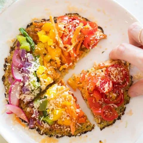 overhead view of a rainbow veggie topped pizza with cauliflower pizza dough and a hand
