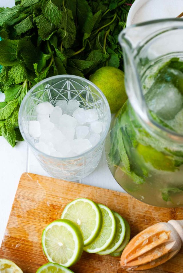 pitcher mojito recipe - a shot of sliced limes with a lemon juicer, a glass full of ice cubes and a pitch full of mojito.